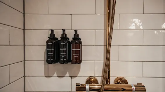 Tre flasker med body wash, shampoo og conditioner monteret på en hvidfliset brusevæg ved siden af et gyldent brusearmatur.