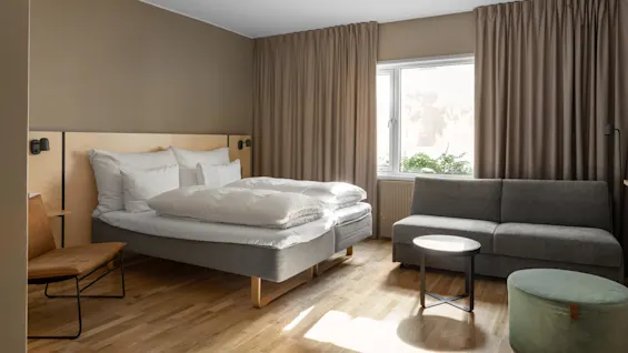 En dubbelsäng står bredvid en liten stol i ett solbelyst rum med trägolv, beige gardiner och en grå soffa under ett fönster.