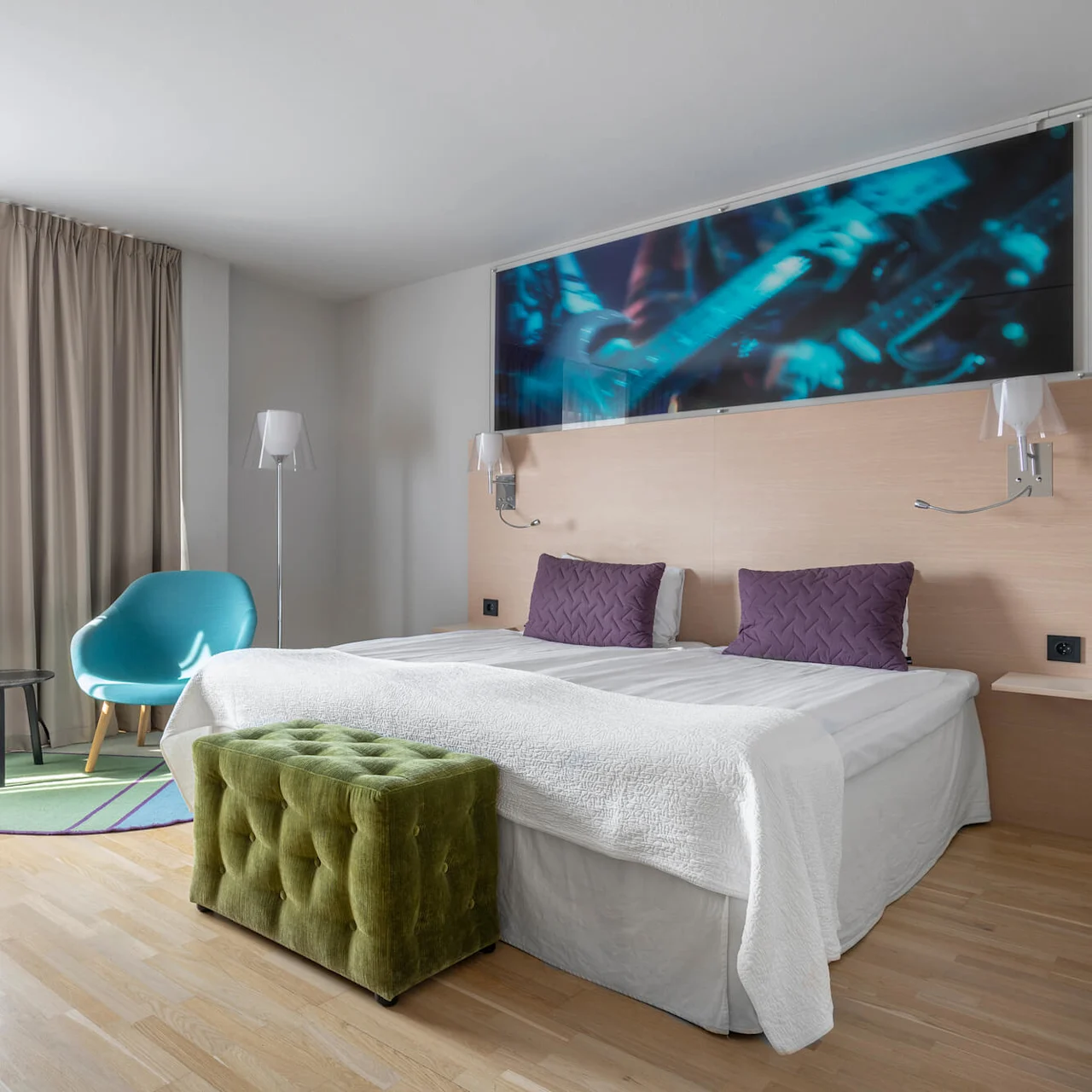 Ett prydligt arrangerat hotellrum med en dubbelsäng med lila kuddar, en grön ottoman vid foten, en blå stol och moderna lampor. Ljusa gardiner filtrerar solljus.