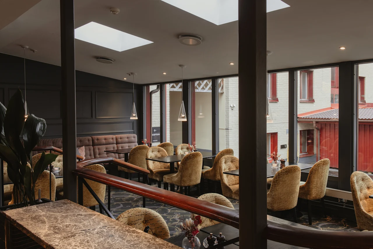 Home Hotel Kung Oscar: A cozy lounge area with comfortable seating, tables, and a reception sign, offering a view outside.