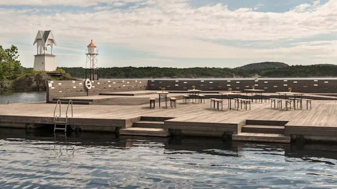 A wooden pier with tables and chairs is set on calm water, surrounded by green hills and buildings, under a partly cloudy sky.