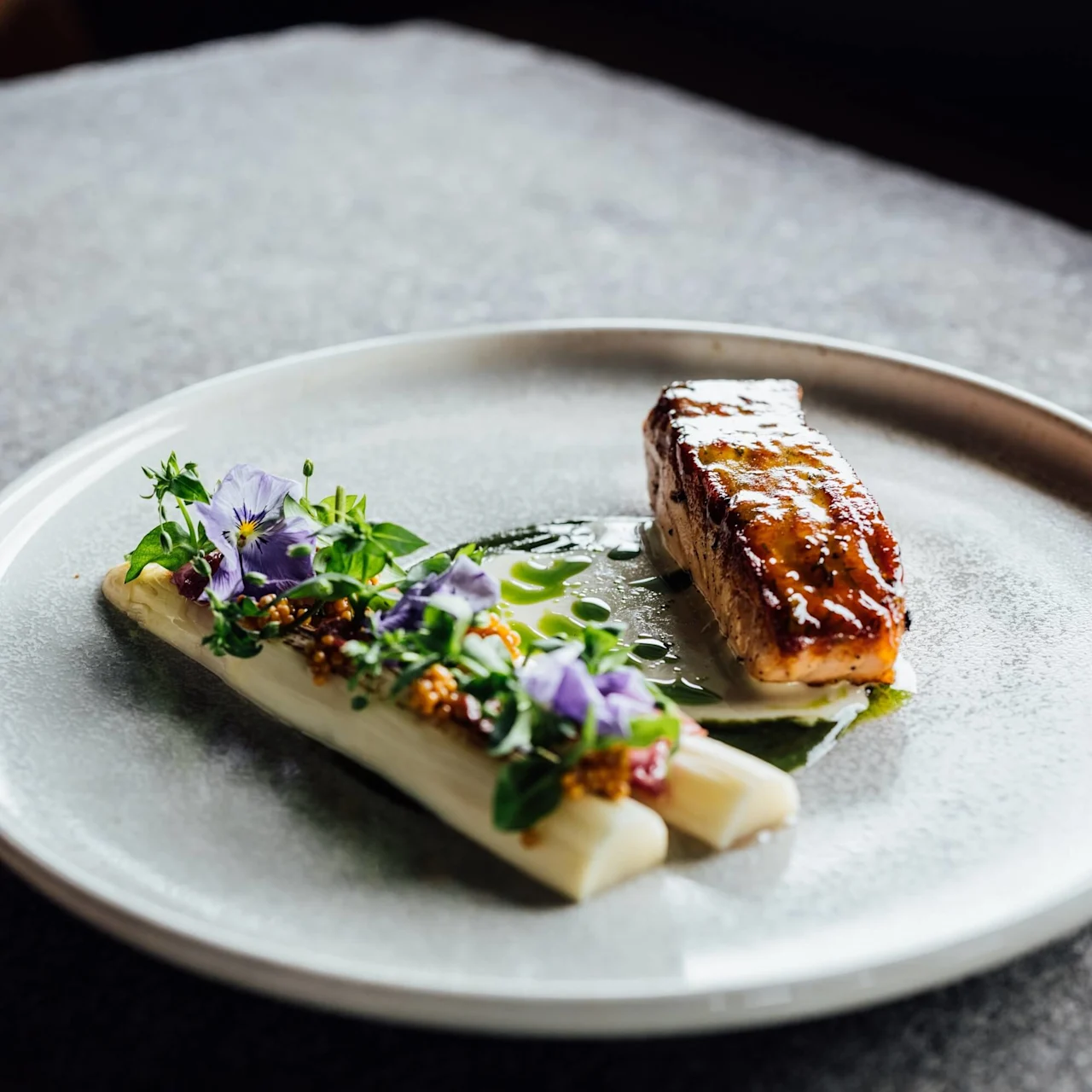 En gourmetret med stegt fisk ved siden af hvid asparges pyntet med urter og lilla blomster, på en tekstureret grå tallerken i dæmpet belysning.