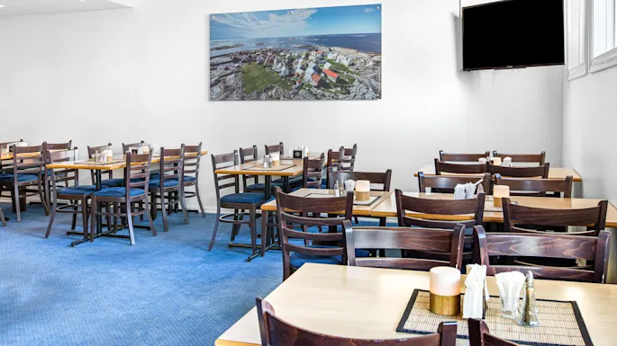Restaurant tables and chairs set for dining, with a large coastal landscape painting on a white wall and a blank television screen nearby. Blue carpet covers the floor.
