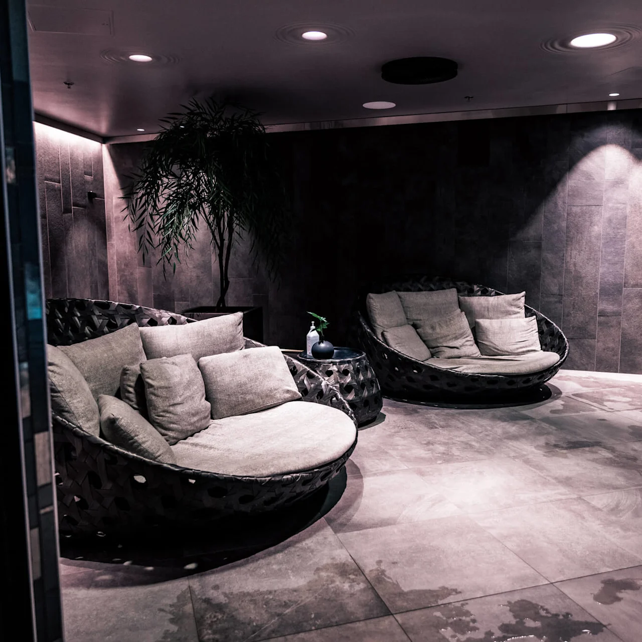 Circular cushioned seating area with pillows rests under soft lighting on a tiled floor, flanked by a small table with a plant. Dark textured walls create a cozy lounge atmosphere.