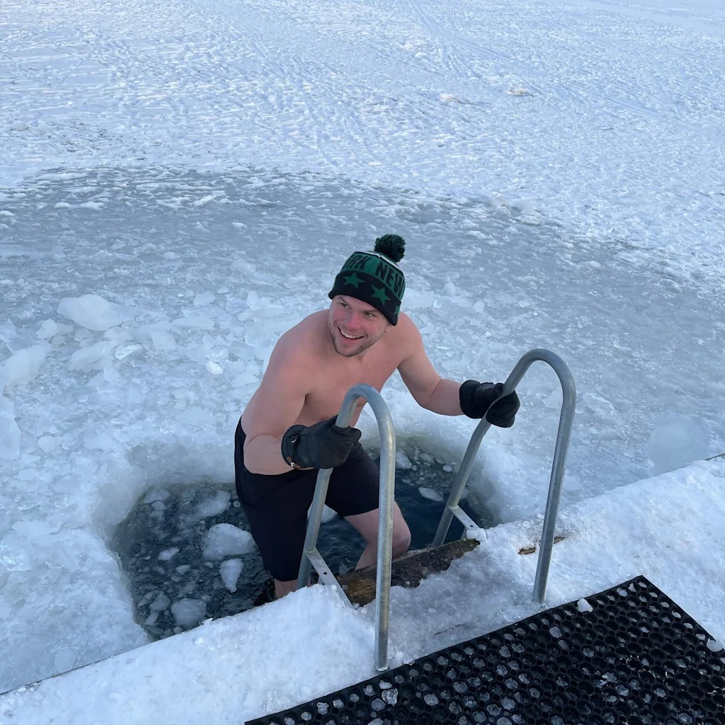 Henkilö hymyilee kiivetessään ylös jääkylvystä jäätyneessä järvessä. Heillä on päässään pipo, jossa lukee NEW. Lumimaisema taustalla.