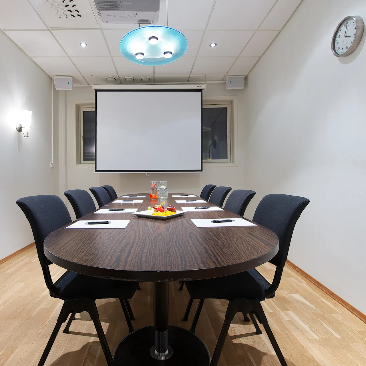 A conference room with a dark oval table set for a meeting, screen on the wall, under artificial lighting.