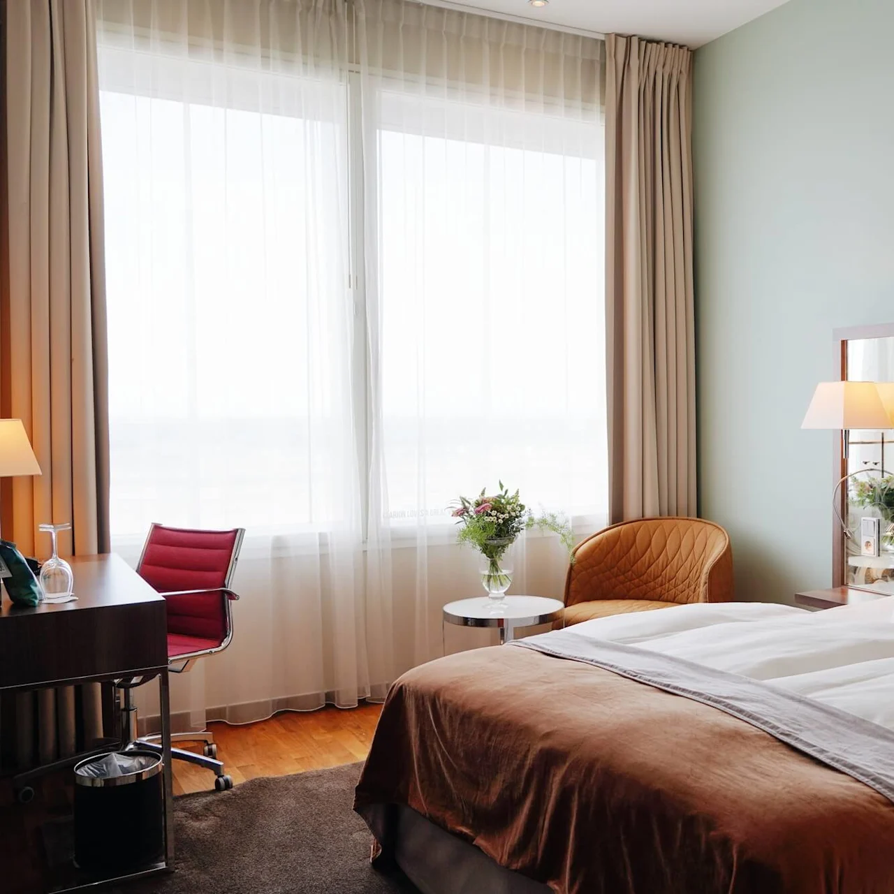 A bed with white linens and brown throw is in a hotel room, accompanied by a desk, chair, lamp, and window with sheer curtains.
