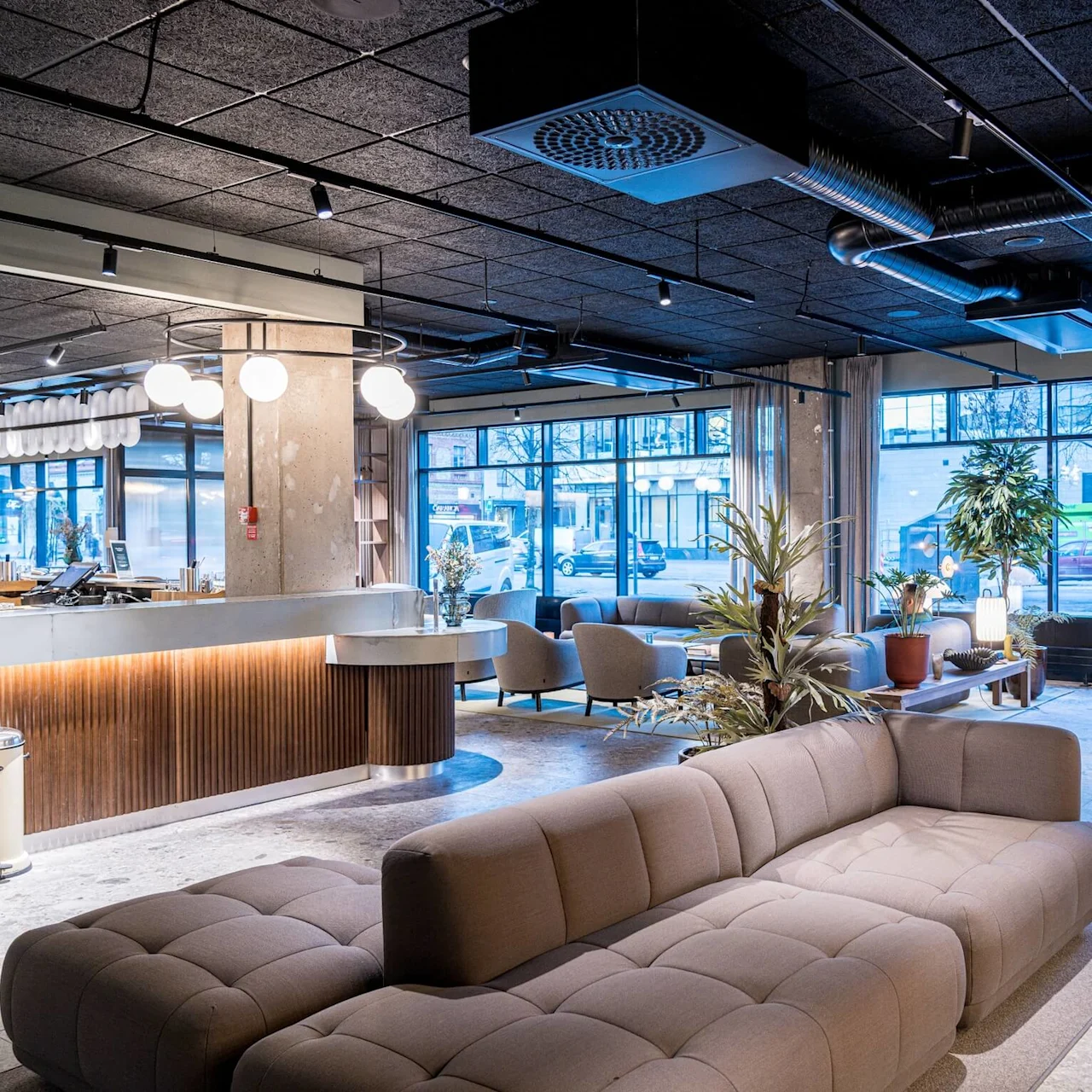 A modern lounge features a large, tufted sofa in a spacious, well-lit room with large windows, surrounded by potted plants and stylish light fixtures.