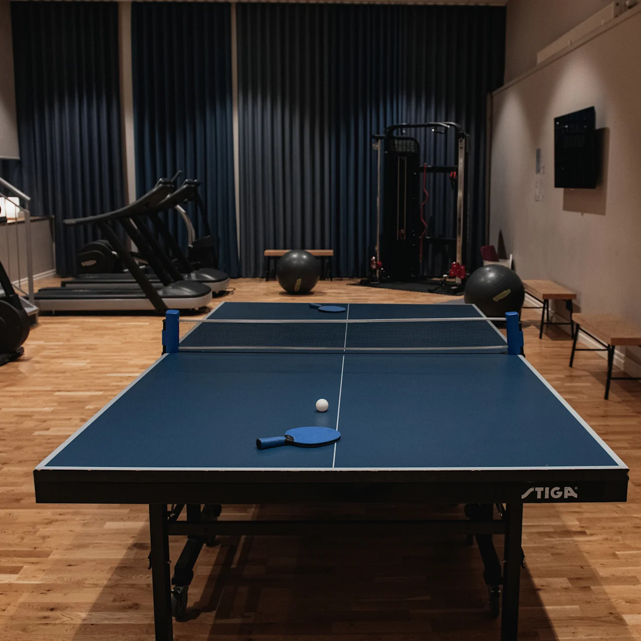 Pingisbord med racket och boll på, i ett gym med träningsutrustning och trägolv; mörka gardiner och bänkar synliga i bakgrunden.