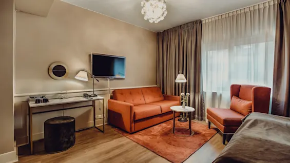 A well-lit hotel room with an orange sofa, coordinating armchair, wall-mounted TV, and desk under soft lighting gives a cozy, elegant ambience.