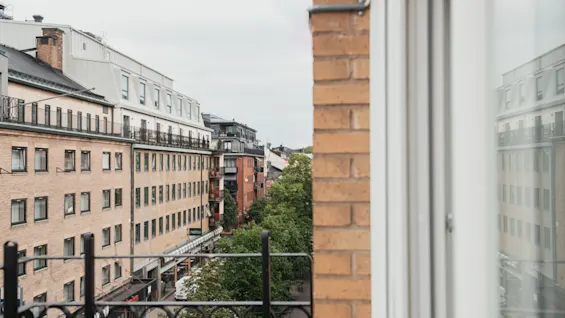 A metal balcony railing overlooks a narrow street between brick buildings in an urban area. Trees line the street below, creating a mix of urban and natural elements.