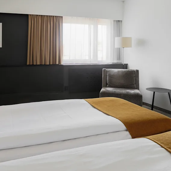 Ett hotellrum har två prydligt bäddade sängar med orangea filtar. I närheten står en grå fåtölj bredvid ett litet runt bord och en lampa. En TV är monterad på den svarta väggen.