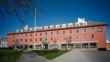En stor, rødorange hotellbygning med mange vinduer under en klarblå himmel, med en asfaltert gangvei og grønt gress foran.