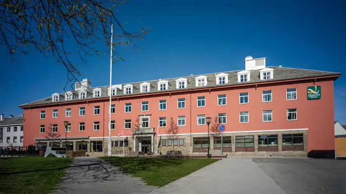 A large, reddish-orange hotel building with many windows under a clear blue sky, with a paved path and green grass in front.