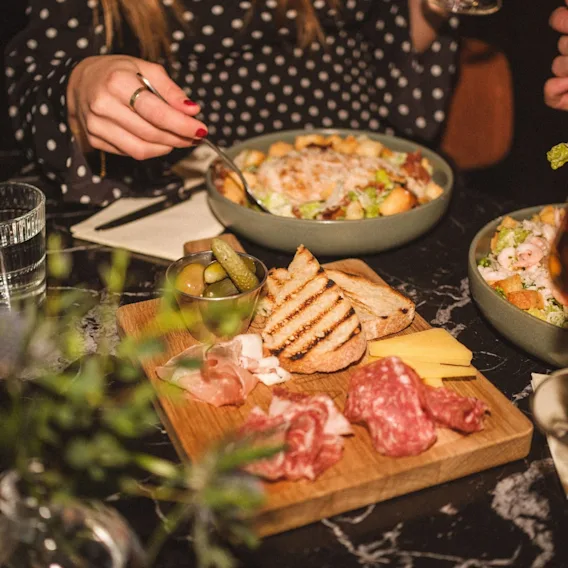 Et spisebord med et trebrett fylt med spekemat, ost, sylteagurk og toast, samt skåler med cæsarsalat.