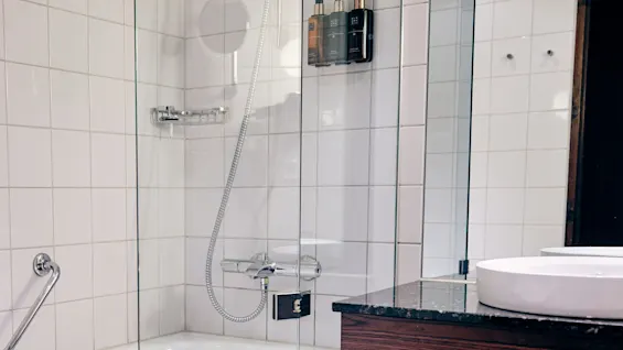 A bathtub with a showerhead is surrounded by white tiled walls and a glass partition. Adjacent is a dark counter with a sink. Several toiletries are mounted on the wall.