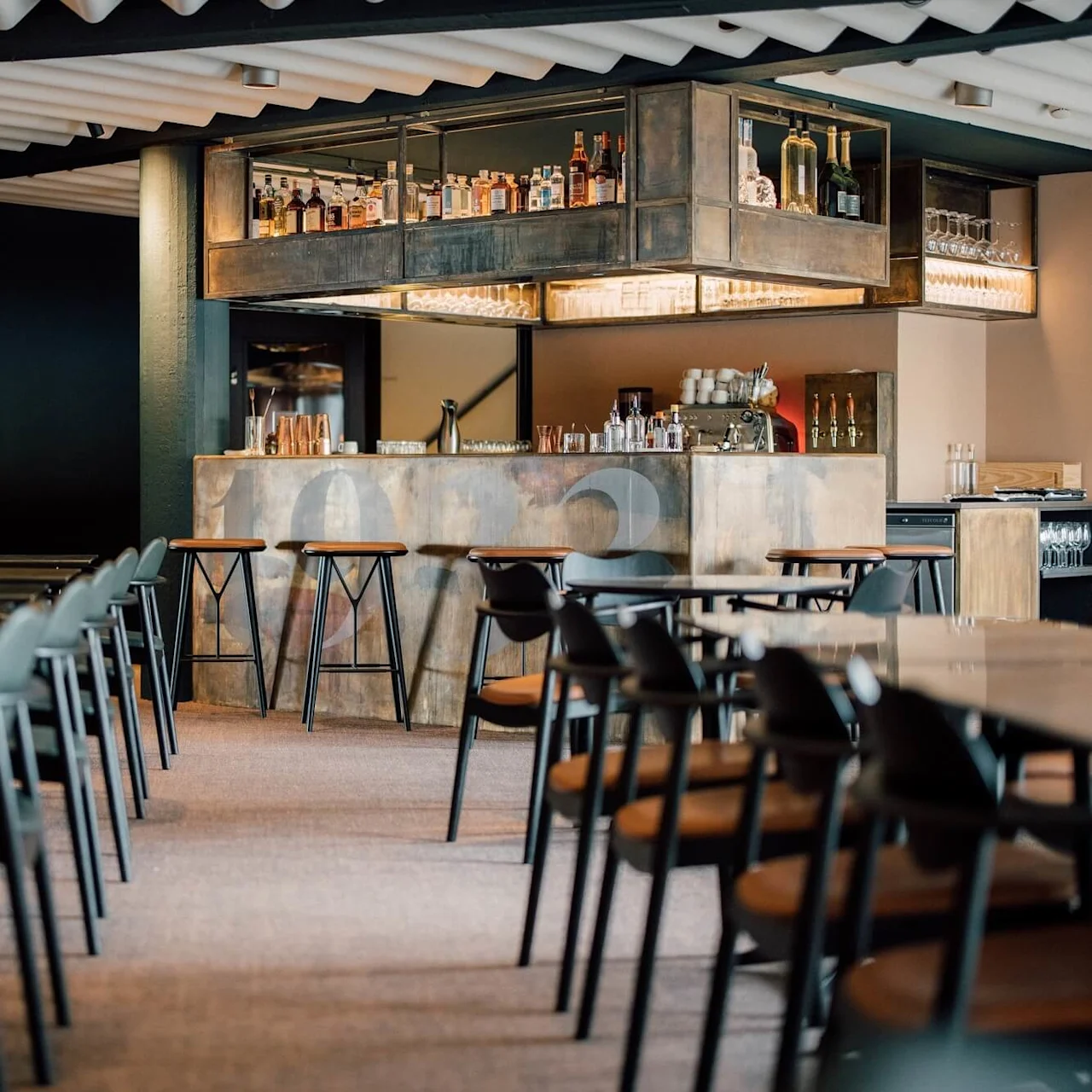 En moderne bar har en elegant skranke med forskellige flasker. Træskamler er stillet foran. Stole og borde fylder det rolige, svagt oplyste rum. Synlig tekst: "restaurant 1988".