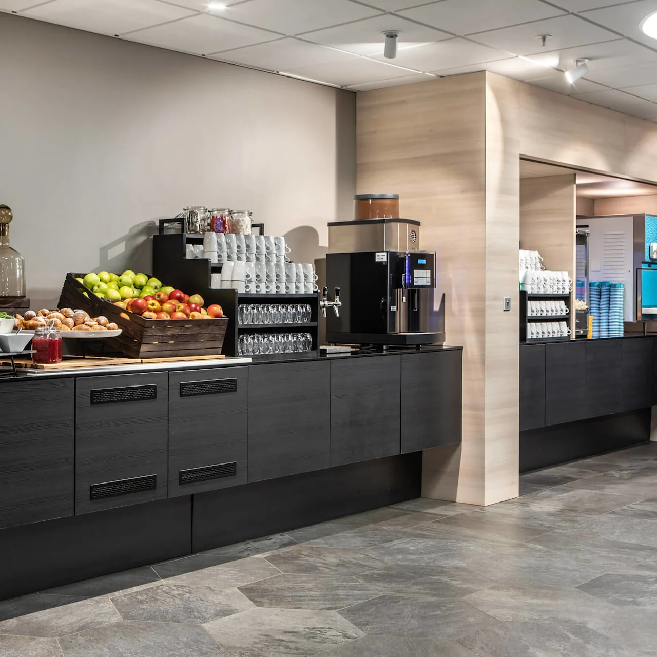 Coffee station featuring various beverages and snacks, set on a modern counter with fruits, pastries, and a coffee machine in a brightly lit room. Wall sign displays "WC" and restroom icons.