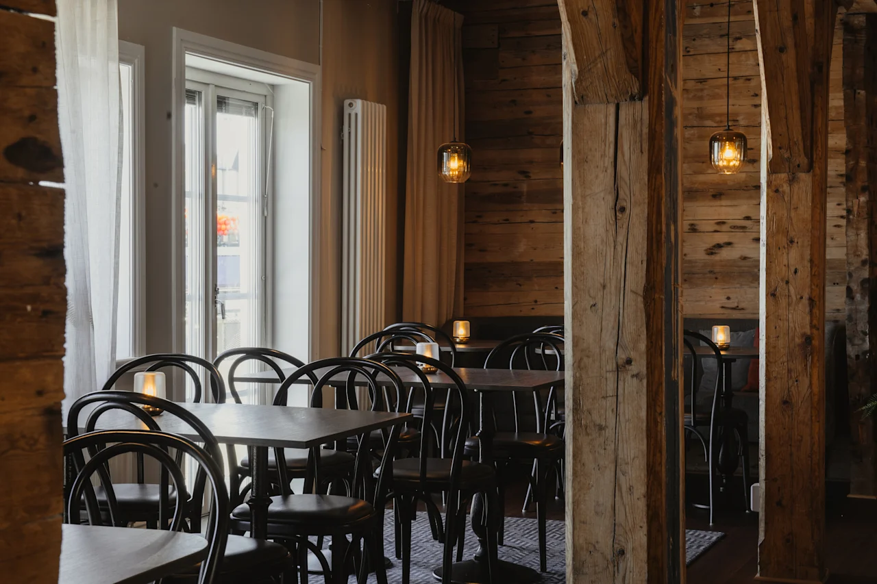 Et hyggeligt spiseområde med træbjælker, borde og stole, oplyst af varme hængende lamper og stearinlys, der skaber en indbydende atmosfære.