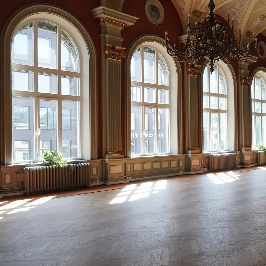 An ornate, empty room bathed in natural light with arched windows, wooden floors, red walls, and decorative chandeliers.