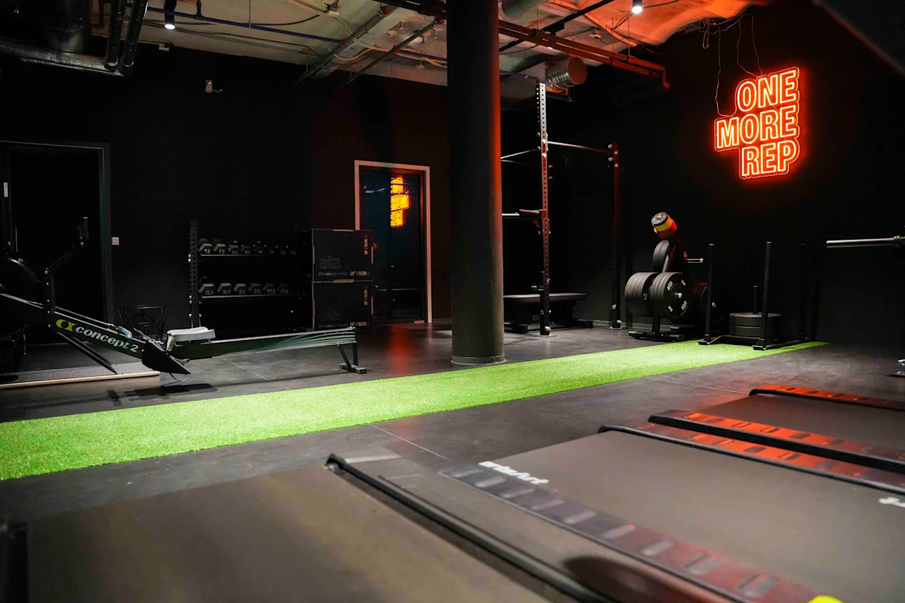 Gyminteriør med træningsudstyr som løbebånd, romaskine og vægte. Neonskilt på væggen med teksten "ONE MORE REP." Mørke vægge og loft med industrielle elementer.