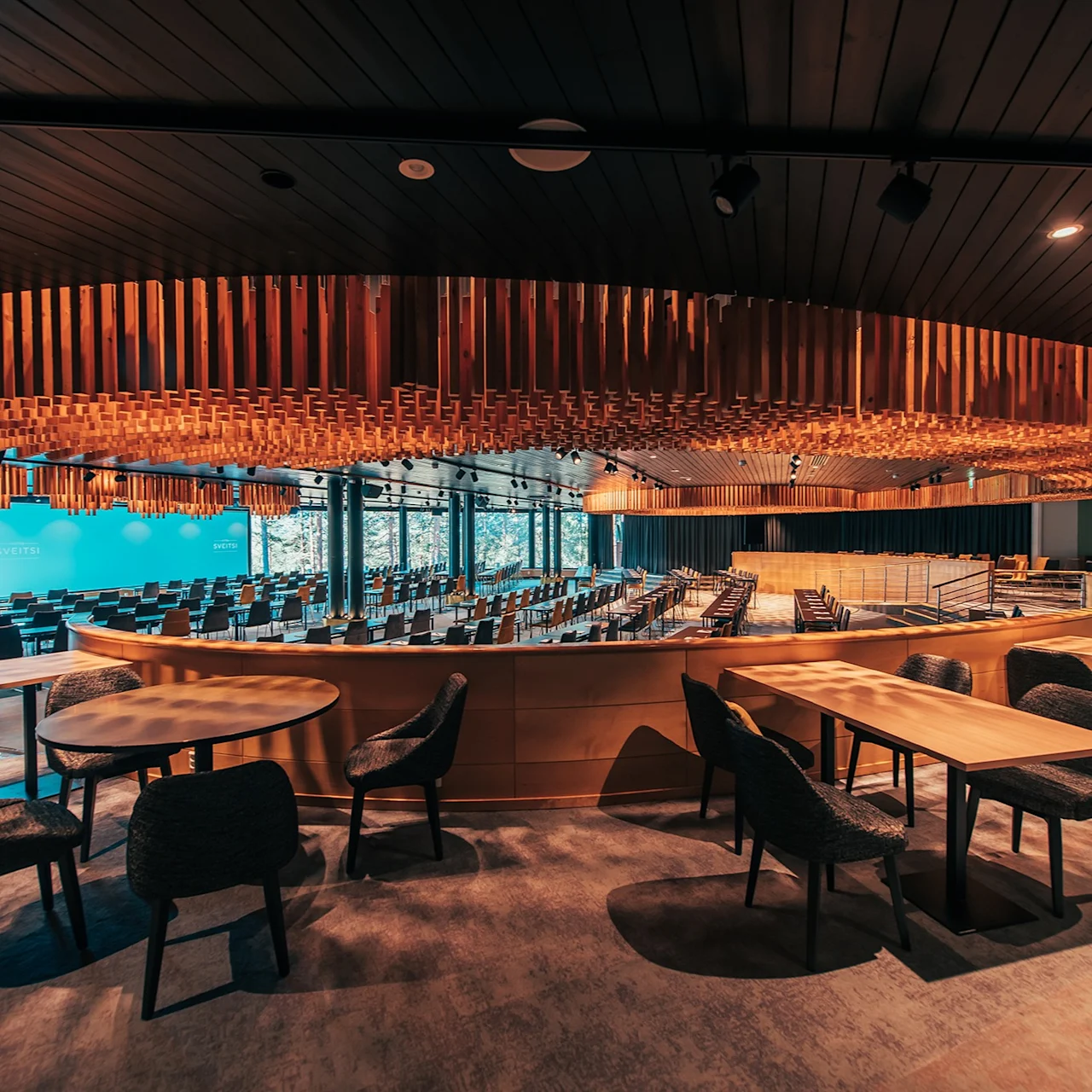 Circular wooden ceiling structure hangs above tables and chairs in a spacious, modern conference room. Rows of seating and large windows are in the background, providing natural light.