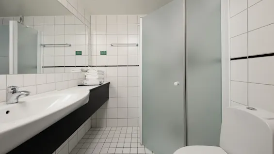 Et rent, moderne badeværelse med hvide fliser, en håndvask, et spejl, en brusekabine og et toilet.