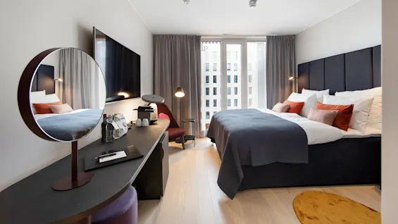 A modern hotel room features a large bed with orange and brown pillows, a wall-mounted TV, and a desk with a mirror. Tall windows have grey curtains. 