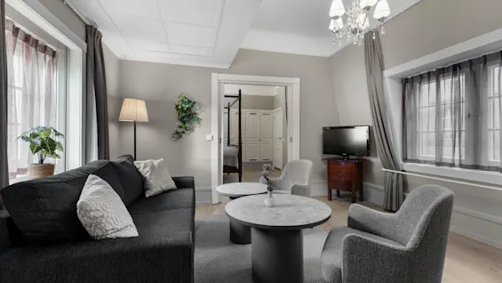 A cozy living room with a dark sofa, two grey armchairs, and coffee tables, leading into a bedroom.