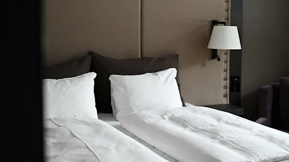 En prydligt bäddad säng med vita kuddar och täcke, placerad mot en beige klädd sänggavel. En väggmonterad lampa finns till höger om sängen.