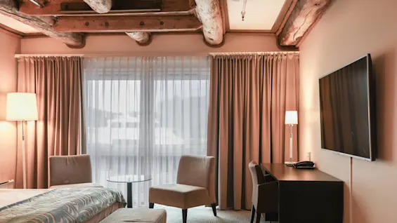 A hotel room with a bed and decorative blanket sits under wooden beams. Adjacent, a desk with a lamp and television faces a window draped with curtains. 