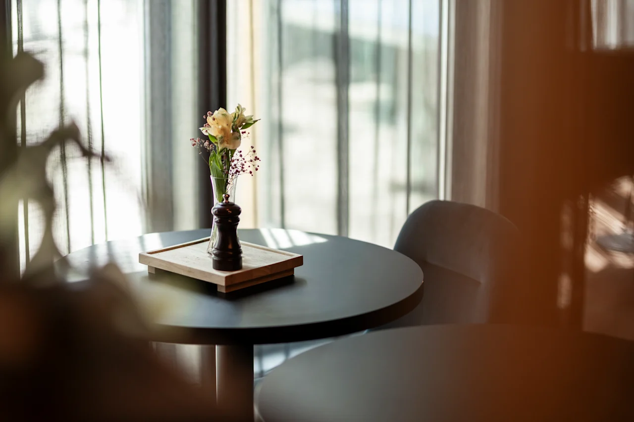 Ett runt bord har en liten vas med blommor och en pepparkvarn på en träbricka i ett mjukt upplyst rum med skira gardiner.