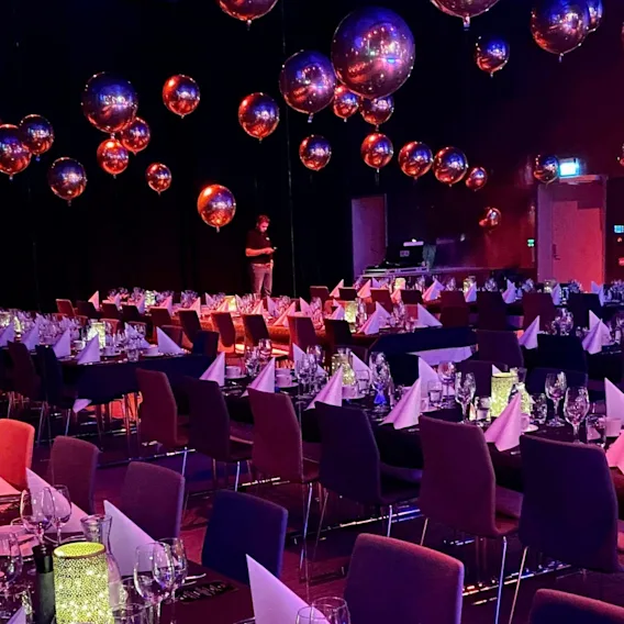 En stor festsal pyntet til fest med dekkede langbord og mange metalliske ballonger som svever under taket i rosa og lilla stemningslys.