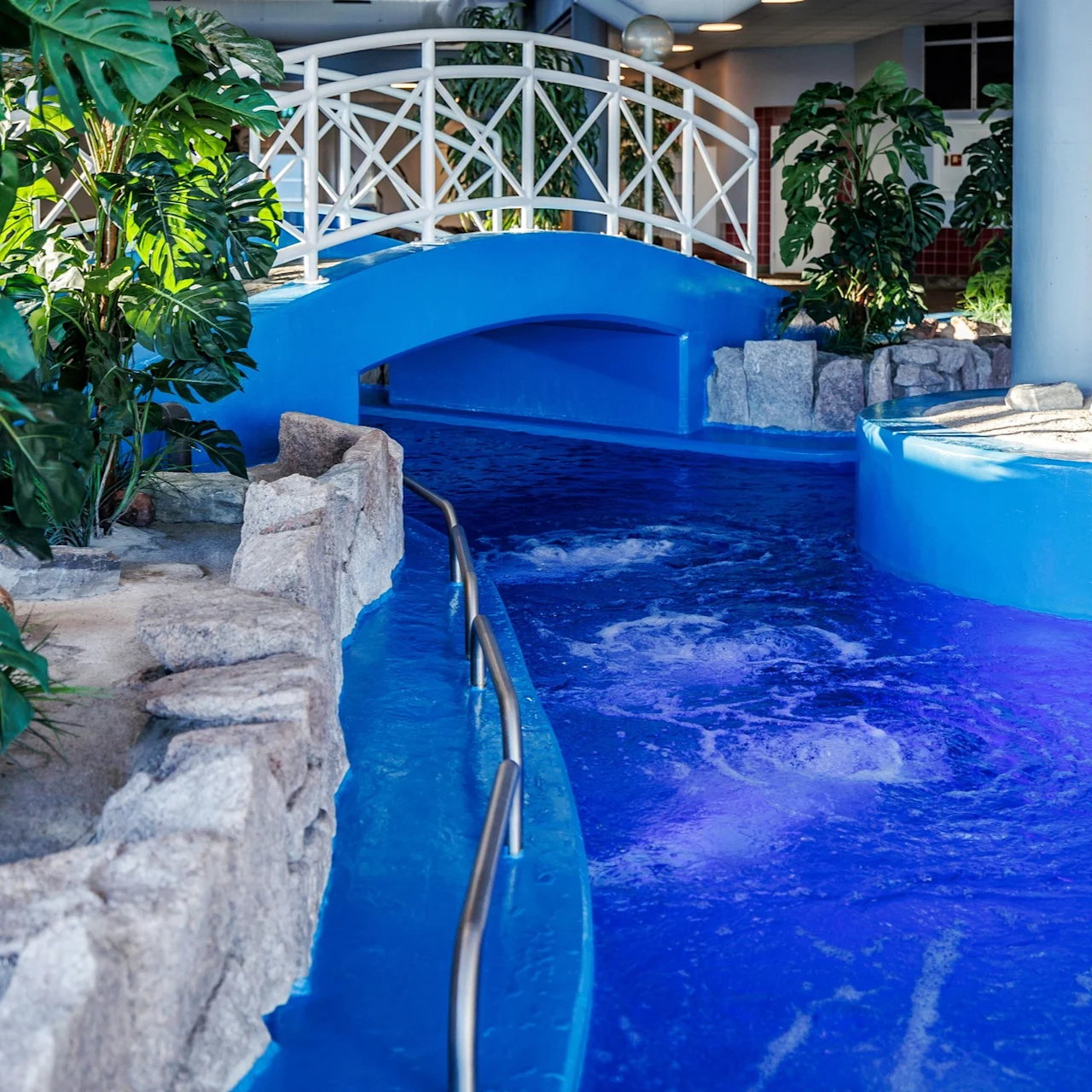 Indoor pool features a small, blue bridge crossing over a flowing water channel, surrounded by artificial rocks and tropical plants, with seating area visible in the background.
