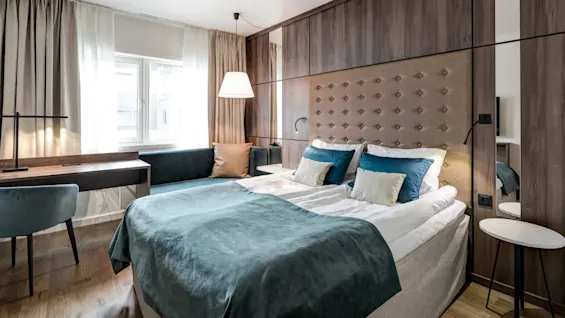 A neatly arranged hotel room features a large bed with white and blue bedding, against a textured wall, accompanied by a window, desk, chair, and soft lighting.