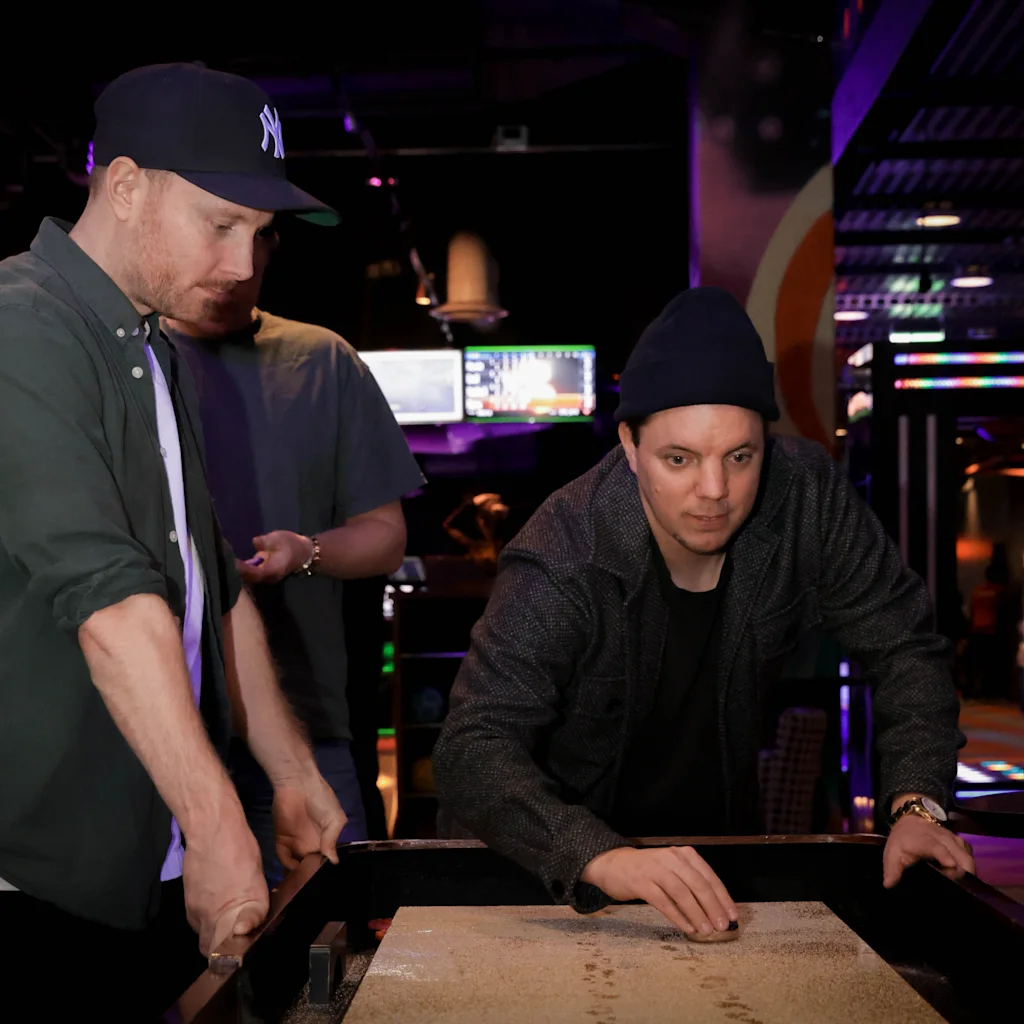 Miehiä pelaamassa shuffleboardia yhdessä tunnelmallisesti valaistussa baarissa.