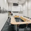 Conference table arranged in a U-shape with water bottles and notepads, surrounded by gray chairs. A projector screen is on the wall, with shelves and wall art nearby.