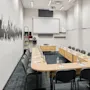 Conference table arranged in a U-shape with water bottles and notepads, surrounded by gray chairs. A projector screen is on the wall, with shelves and wall art nearby.