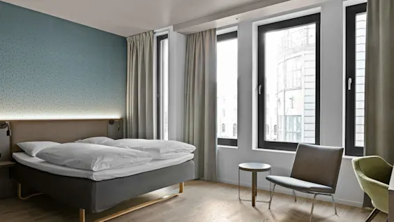 A modern hotel room features a neatly made bed, with two pillows, beside large windows. A small round table and chair sit nearby, illuminated by natural light.