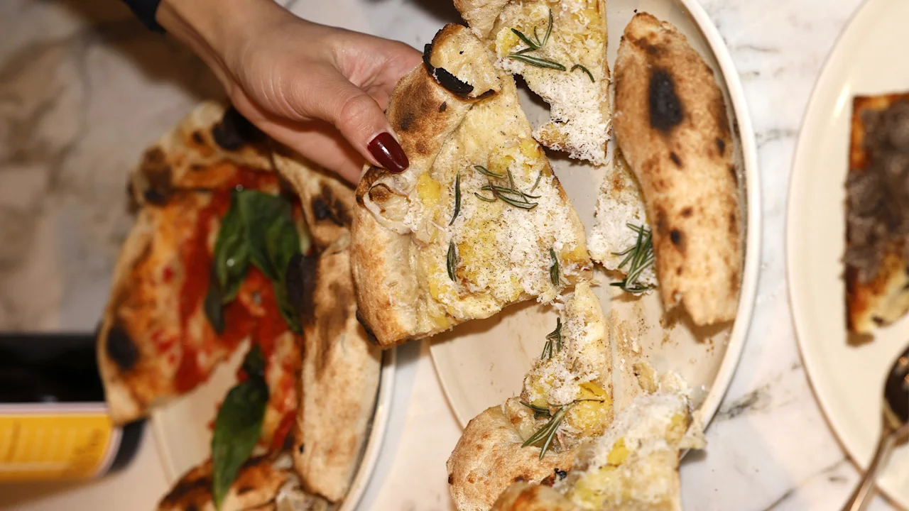 En hånd løfter et pizzastykke med rosmarin fra en tallerken, sammen med en annen pizza med tomat og basilikum. Tallerkener hviler på en marmoroverflate.