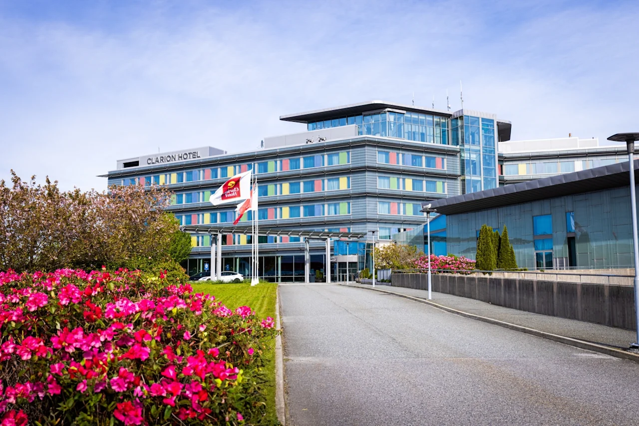 Et moderne fleretasjes hotell med påskriften "CLARION HOTEL" står blant velstelte hager med livlige rosa blomster. Scenen er lys under en klar blå himmel. Et flagg i nærheten viser hotellets logo.