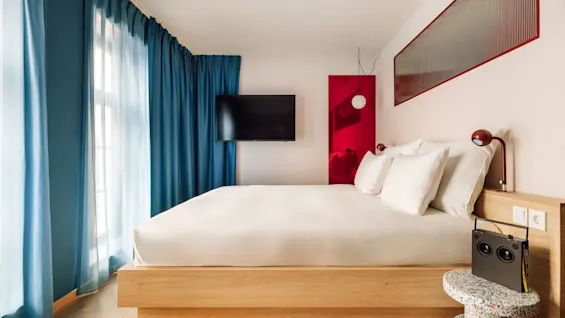 A vibrant hobo oslo hotel room featuring a cozy bed, bright blue curtains, and a stylish red wall accent.