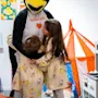 En karakter i et pingvindrakt klemmer to barn i et koselig, lekent rom med fargerik kunst og et lite leketelt. Barna har matchende klær, deler et øyeblikk av glede og varme.