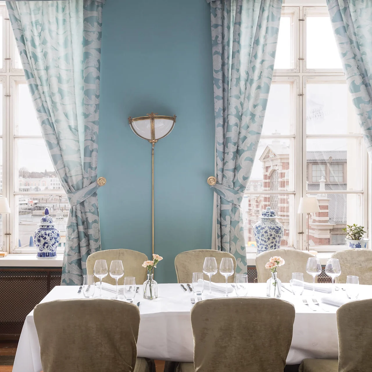 A white table set with glasses and flowers in vases, surrounded by upholstered chairs, stands near blue-patterned curtains and windows in a well-lit dining room, overlooking buildings outside.
