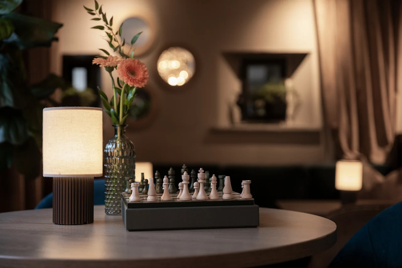 A cozy corner at Home Hotel Grand Bodø with a chess set ready for a game, illuminated by a warm lamp and fresh flowers.