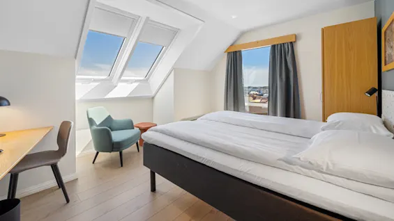 A well-lit attic bedroom with a large bed, a skylight, and modern furniture, overlooking an urban landscape.