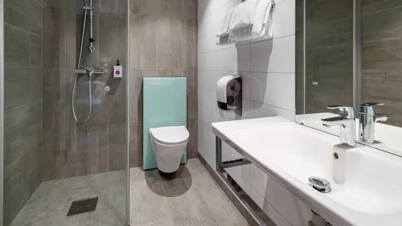 Shower stall stands enclosed by glass in a modern bathroom with toilet, sink, and towels on a rack. Neutral tiled walls and floor with a large mirror create a sleek ambiance. No text present.