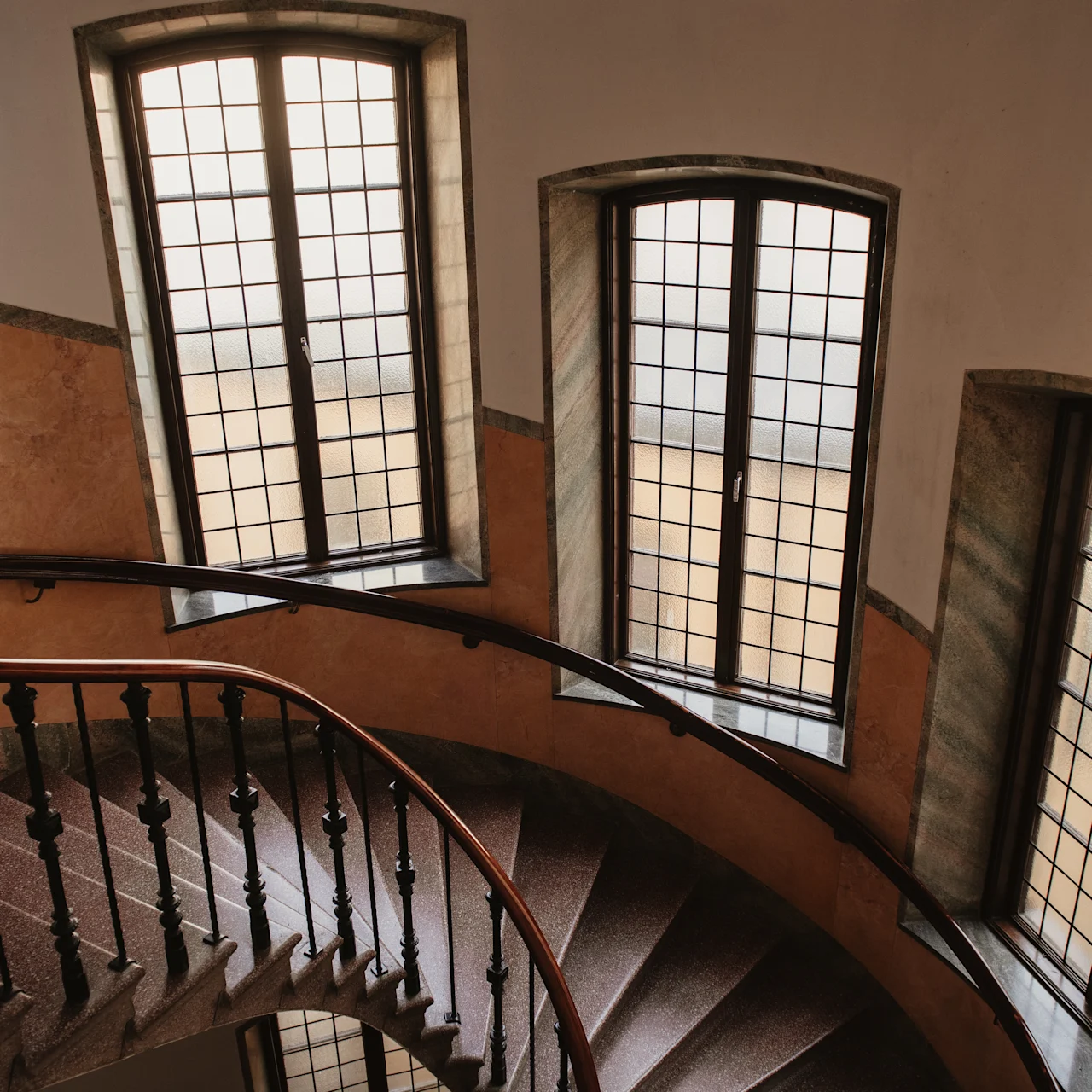 Spiraltrappa med träbalustrad kröker nedåt bredvid välvda fönster, som kastar mjukt ljus i ett smalt, inneslutet utrymme.