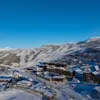 En flygbild över en snötäckt skidort med byggnader inbäddade bland bergen och synliga skidbackar under en klarblå himmel.