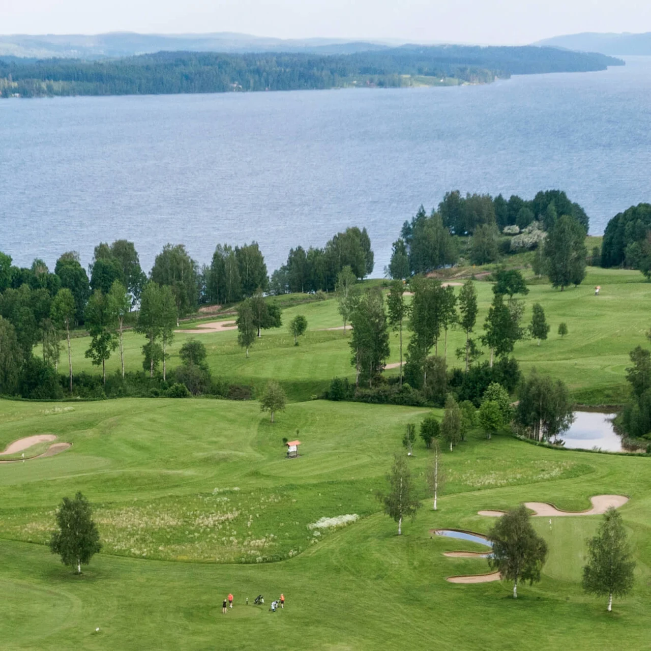 Golfbane med spillere på grønt gress, omgitt av trær, og med en stor innsjø i bakgrunnen.